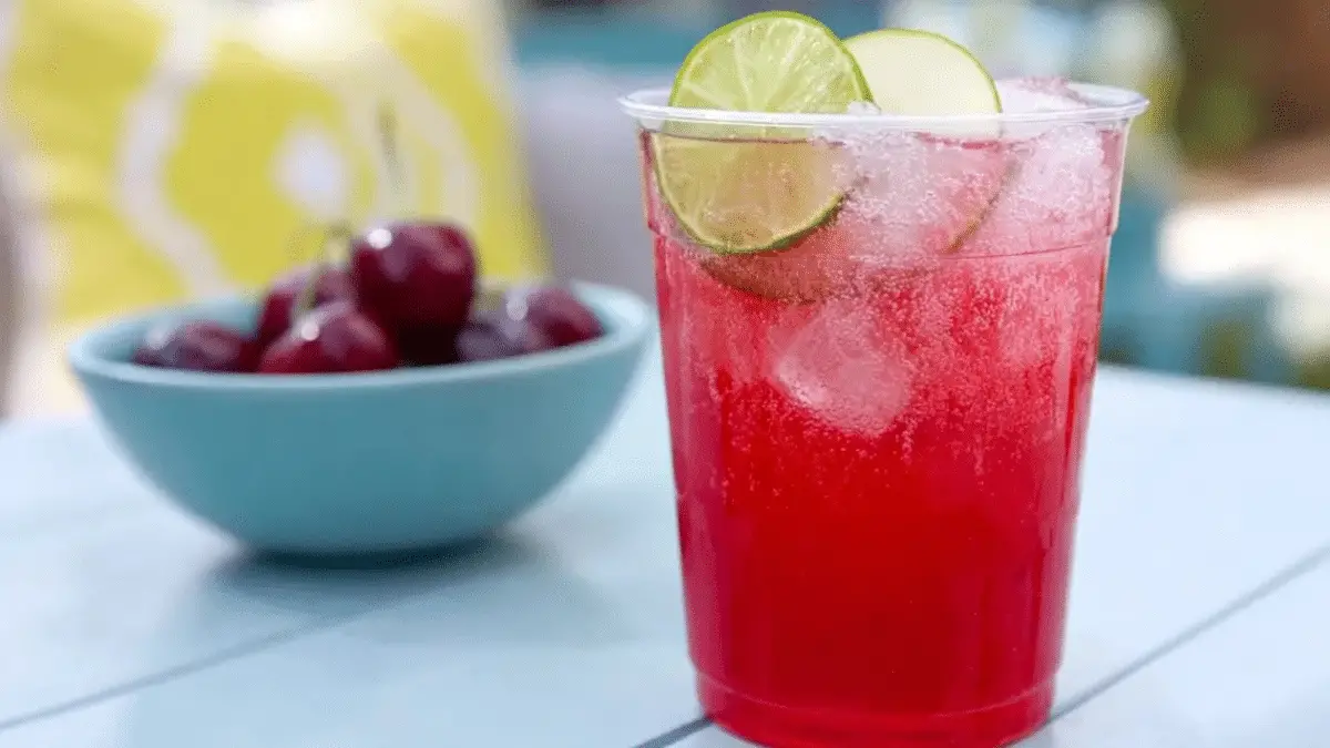 Refreshing cherry lime rickey served in a clear plastic cup with ice and lime slices, next to a bowl of fresh cherries.
